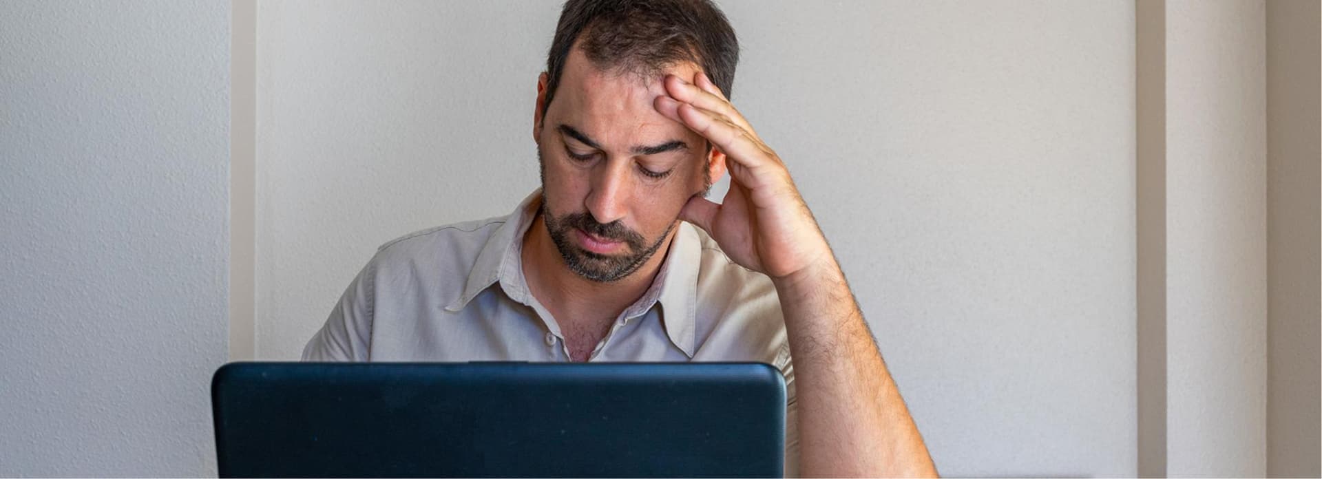 A bearded man looks stressed while sitting in front of a laptop, resting his head on one hand, in a brightly lit room.