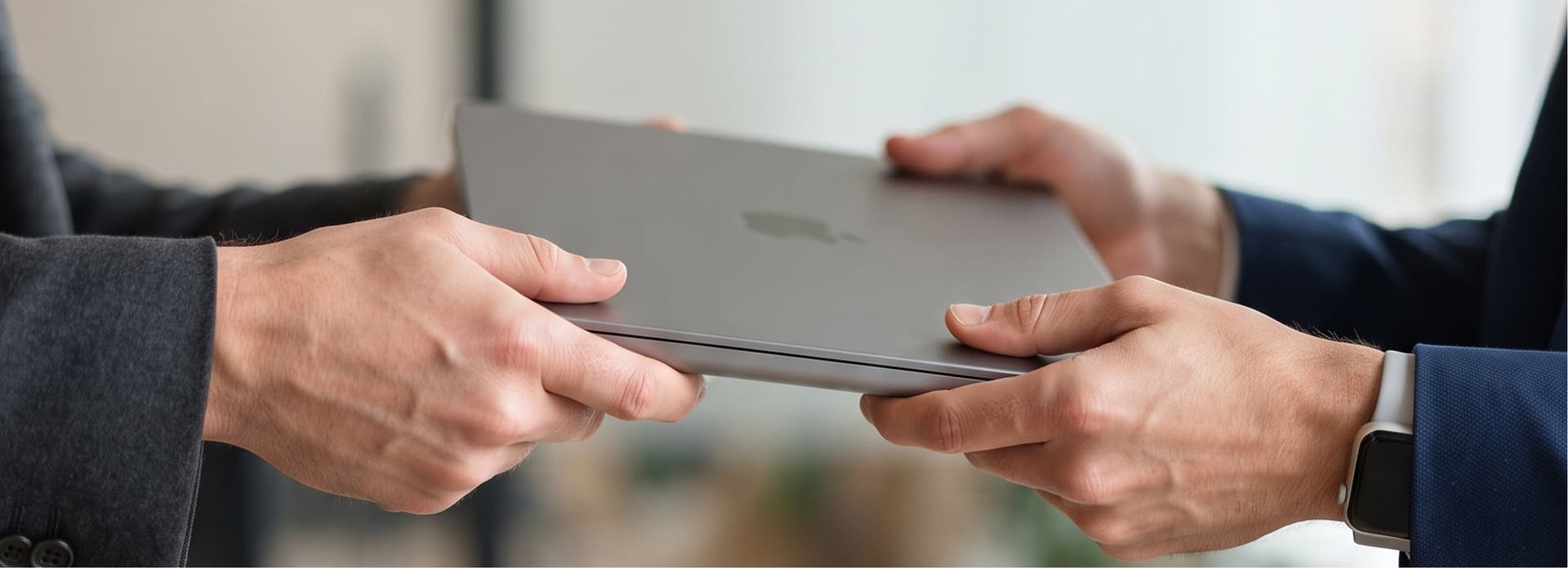 Two people exchanging a closed laptop, one wearing a smartwatch, indoors with blurred background.