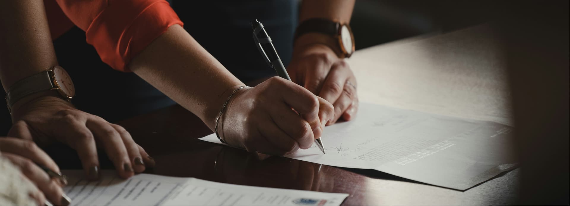 Hands of two people on a desk signing documents, one holding a pen. Both wear watches and one has a red sleeve.